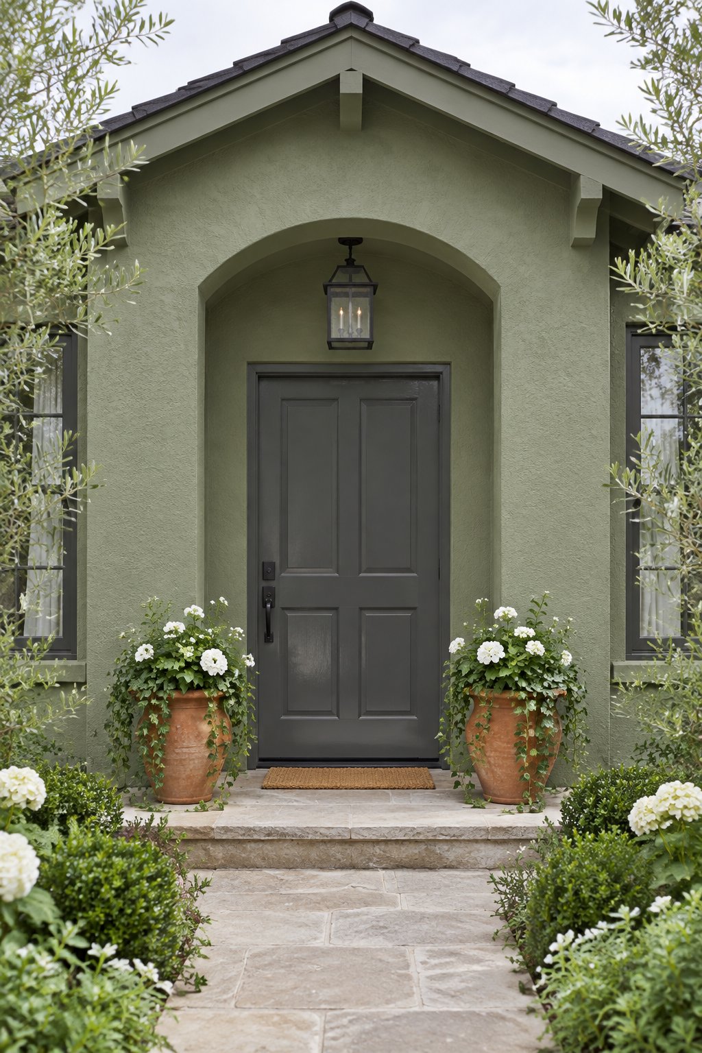 A front entrance with sage green walls, a charcoal gray door, and terracotta pots of ivy and white flowers.